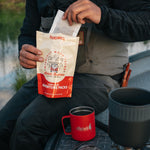 Person pouring a Kaladi Adventure Pack single-serve coffee into a red camp mug outdoors
