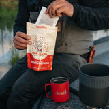 Person pouring a Kaladi Adventure Pack single-serve coffee into a red camp mug outdoors