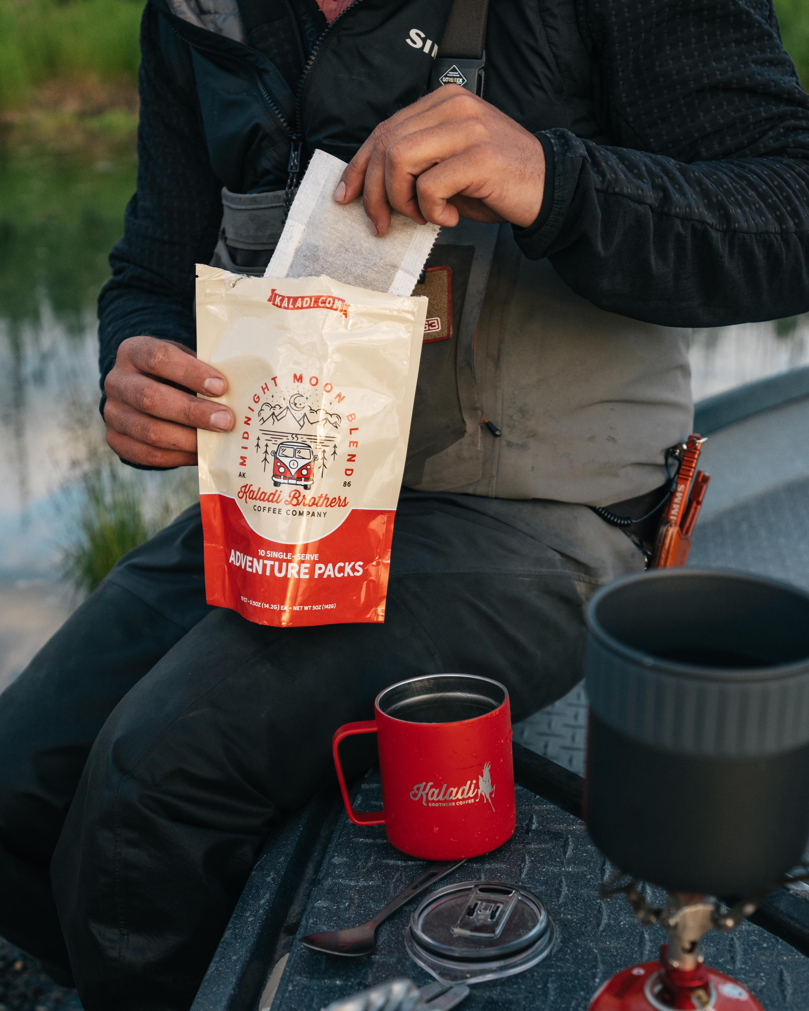 Person pouring a Kaladi Adventure Pack single-serve coffee into a red camp mug outdoors