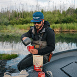 Man brewing Kaladi coffee on a boat beside an Alaska river