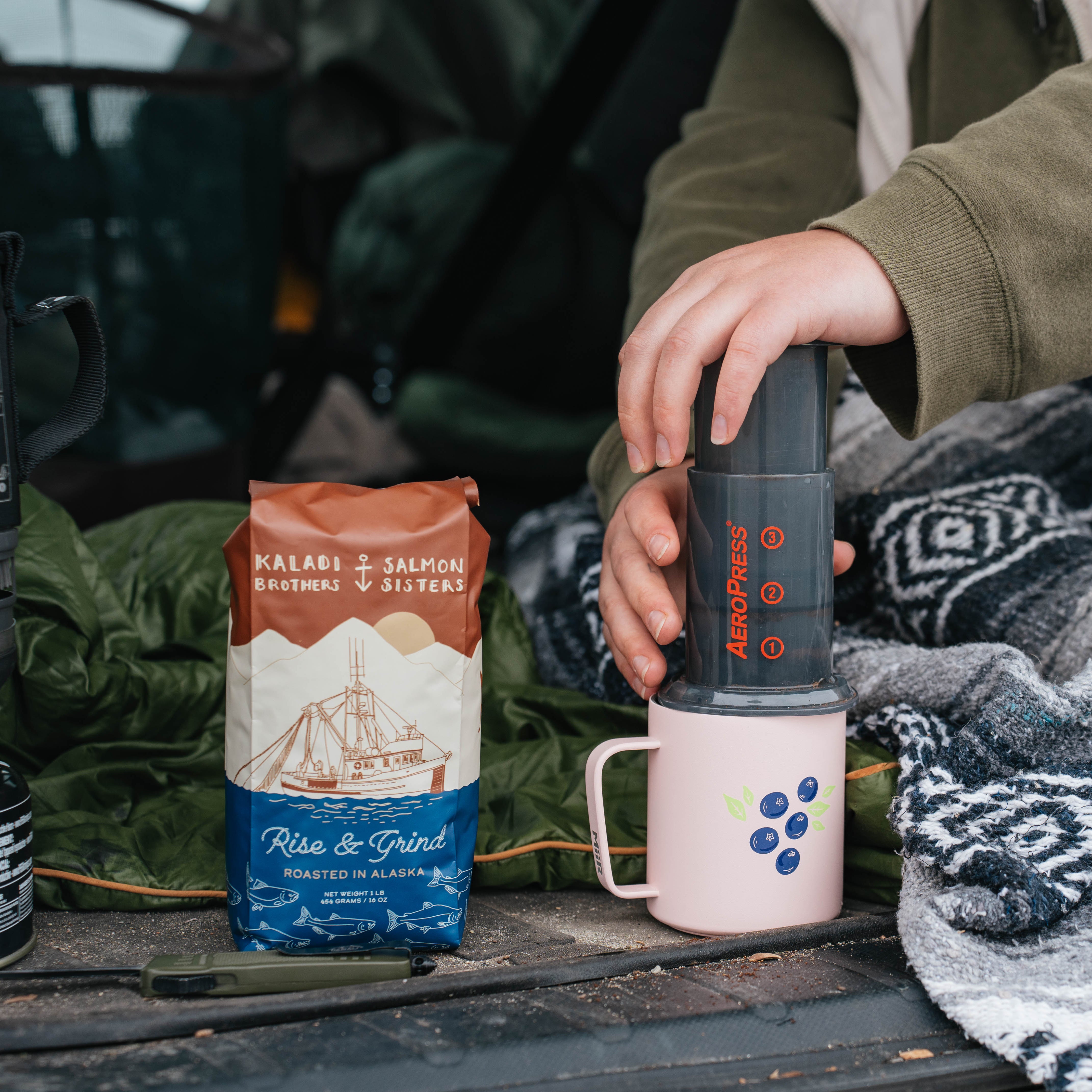 Person preparing a hot beverage with a coffee maker and coffee bags on a camping mat.