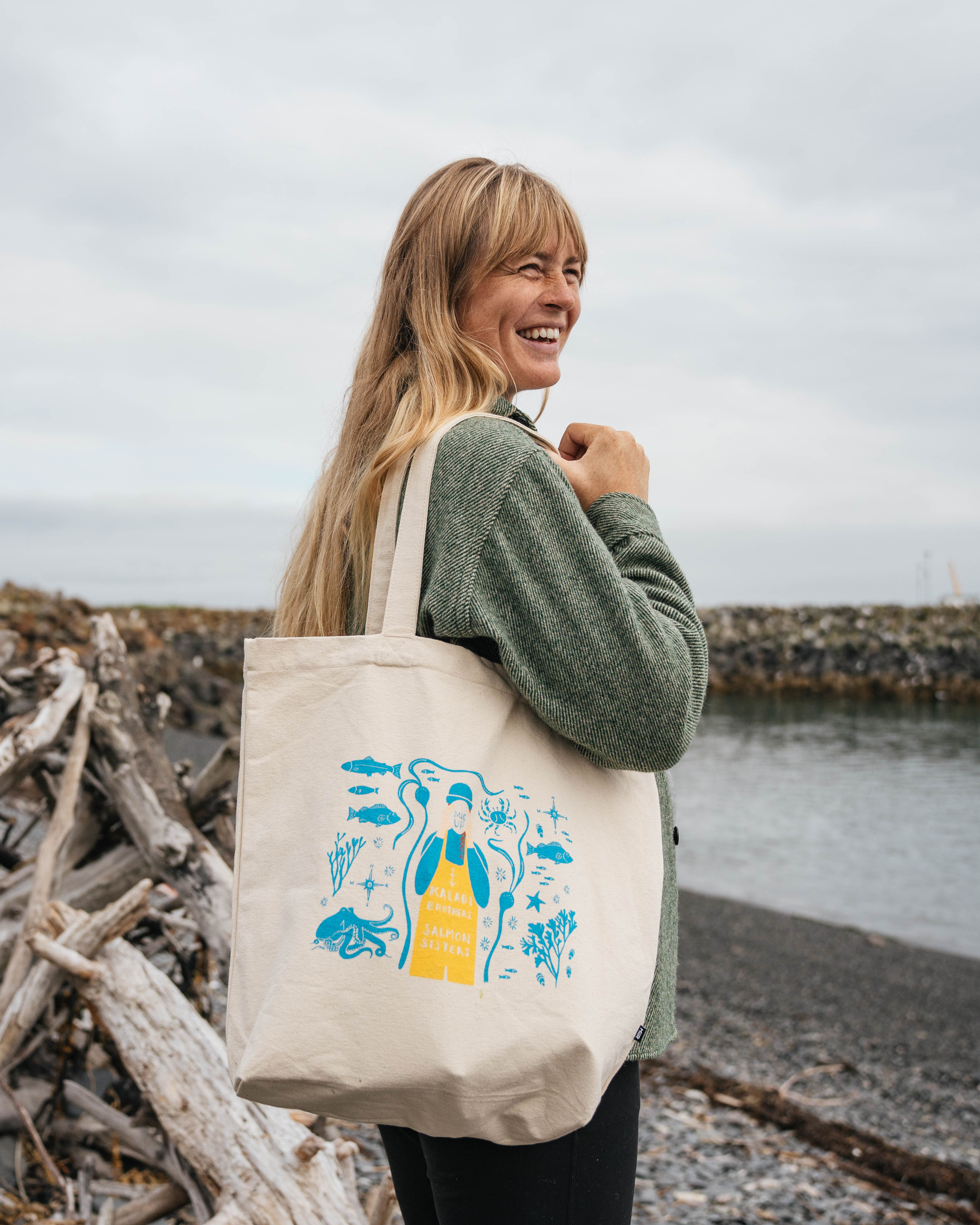 Woman on a rocky beach carrying the Salmon Sisters canvas tote