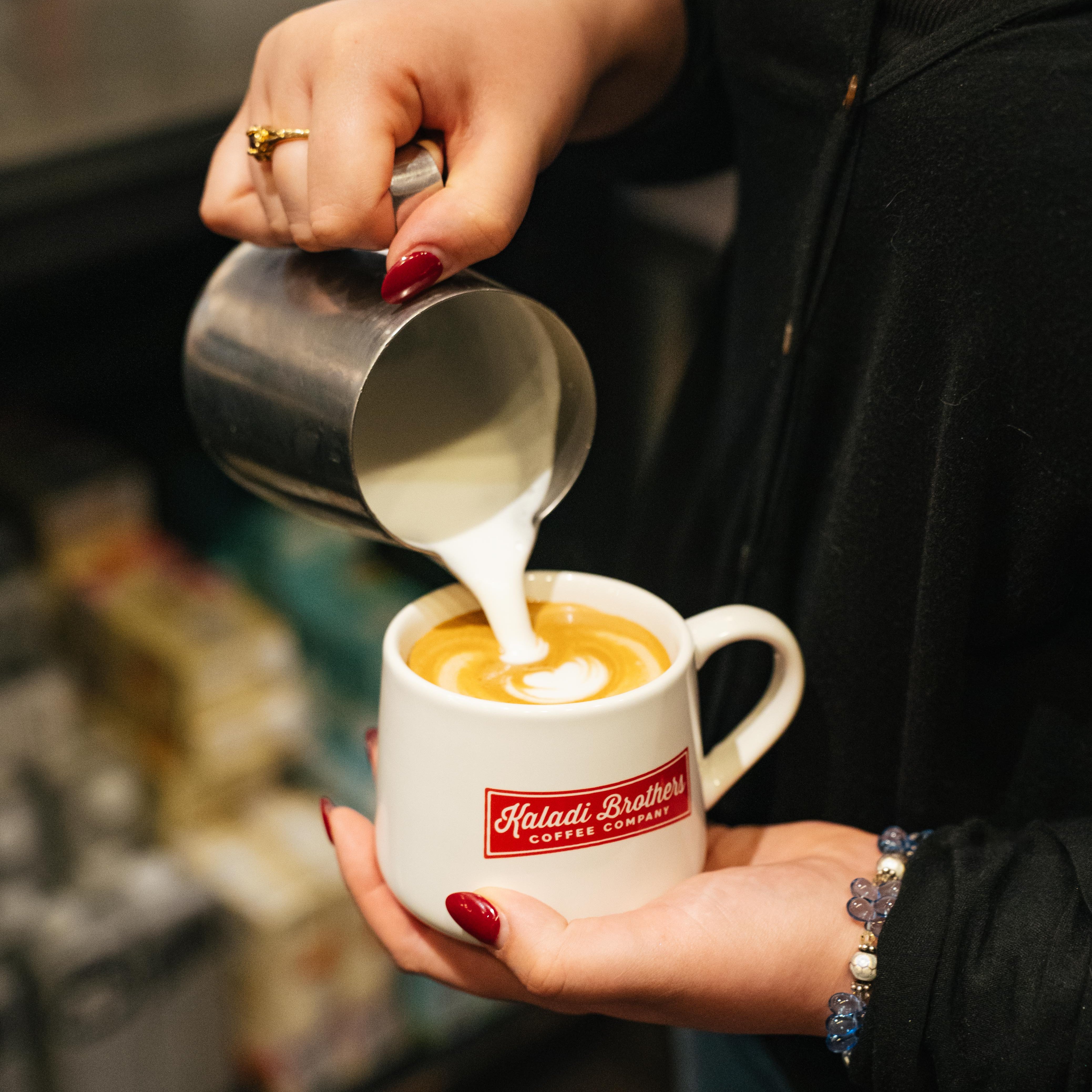 A person pouring steamed milk from a stainless steel pitcher into a white coffee cup with a red and white 'Haisati Brothers Coffee Company' logo, creating latte art. The person is wearing a black top and has red nail polish.