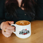 A person holding a white ceramic coffee mug with a black, red, and white mountain truck graphic, sitting on a wooden table in a cafe setting.