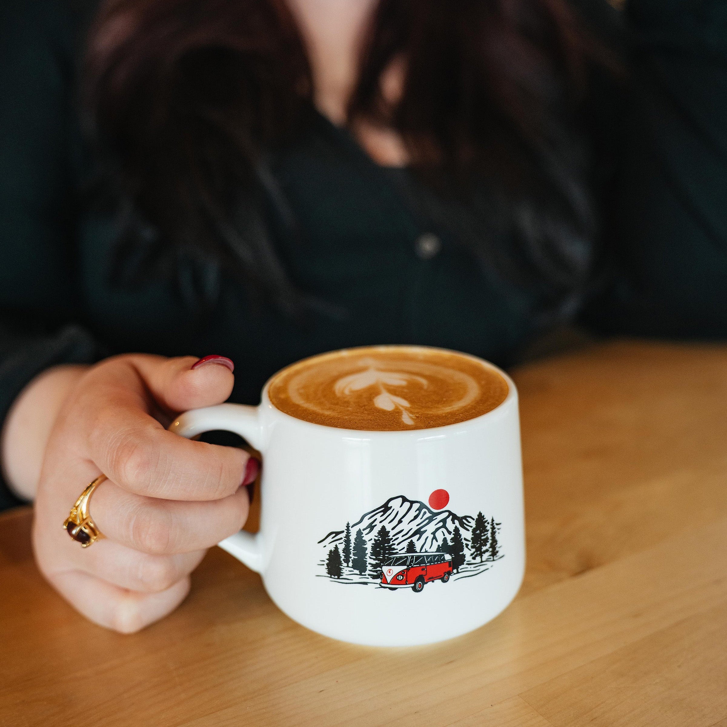 A person holding a white ceramic coffee mug with a black, red, and white mountain truck graphic, sitting on a wooden table in a cafe setting.