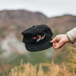 Hand holding the black Coffee Cherry Field Trip hat against a hillside
