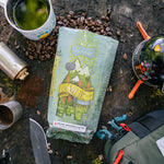 Coffee package labeled 'Big Wild Life' on a wooden surface with coffee beans and camping equipment.