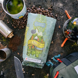 Coffee package labeled 'Big Wild Life' on a wooden surface with coffee beans and camping equipment.