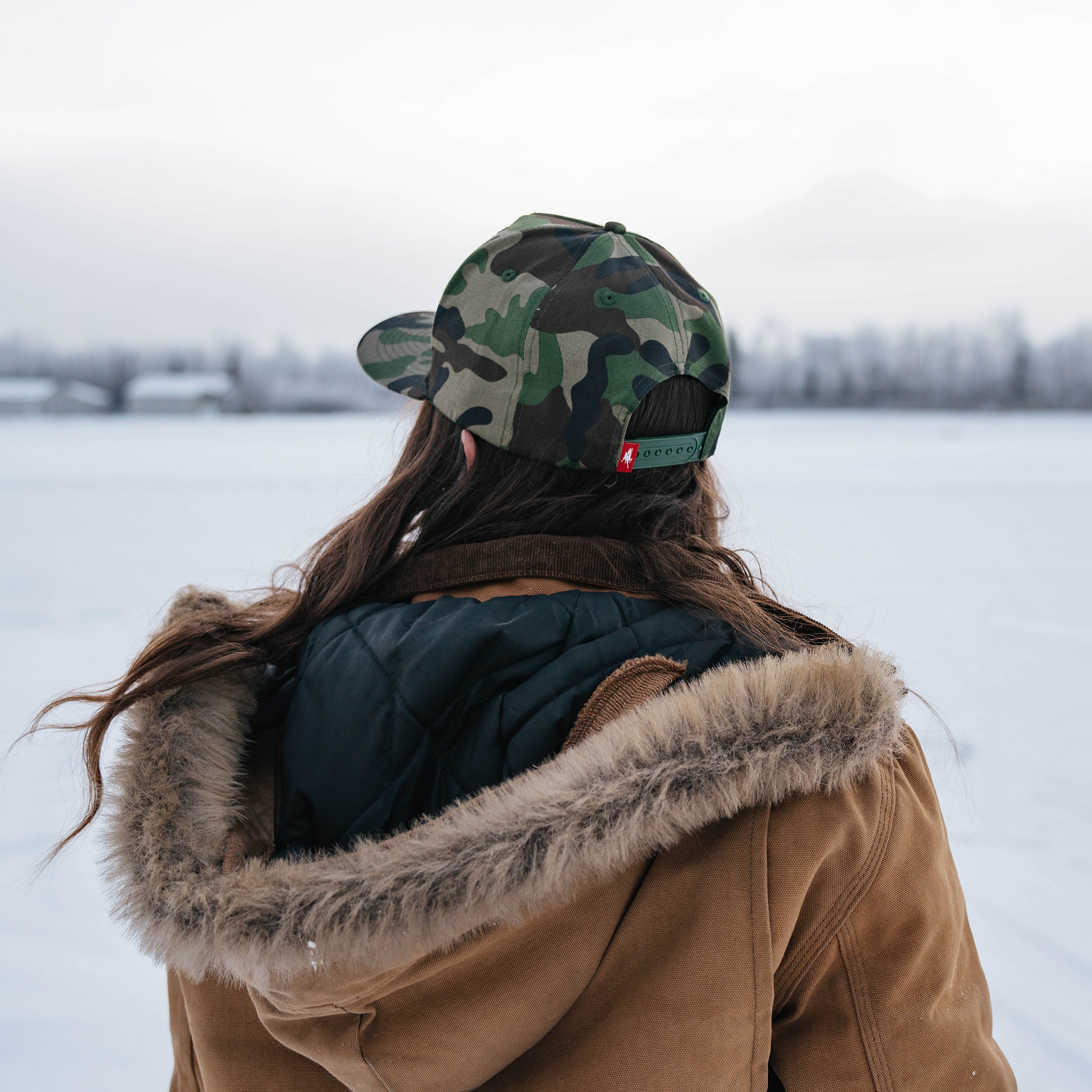 Woman in a fur-lined coat and Kaladi camo hat looking toward snowy mountains