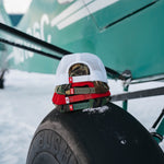 Kaladi camo and white trucker hats stacked on a bush plane tire in the snow