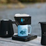 Coffee brewing setup with a blue mug, black filter holder, and black thermos on a wooden surface.