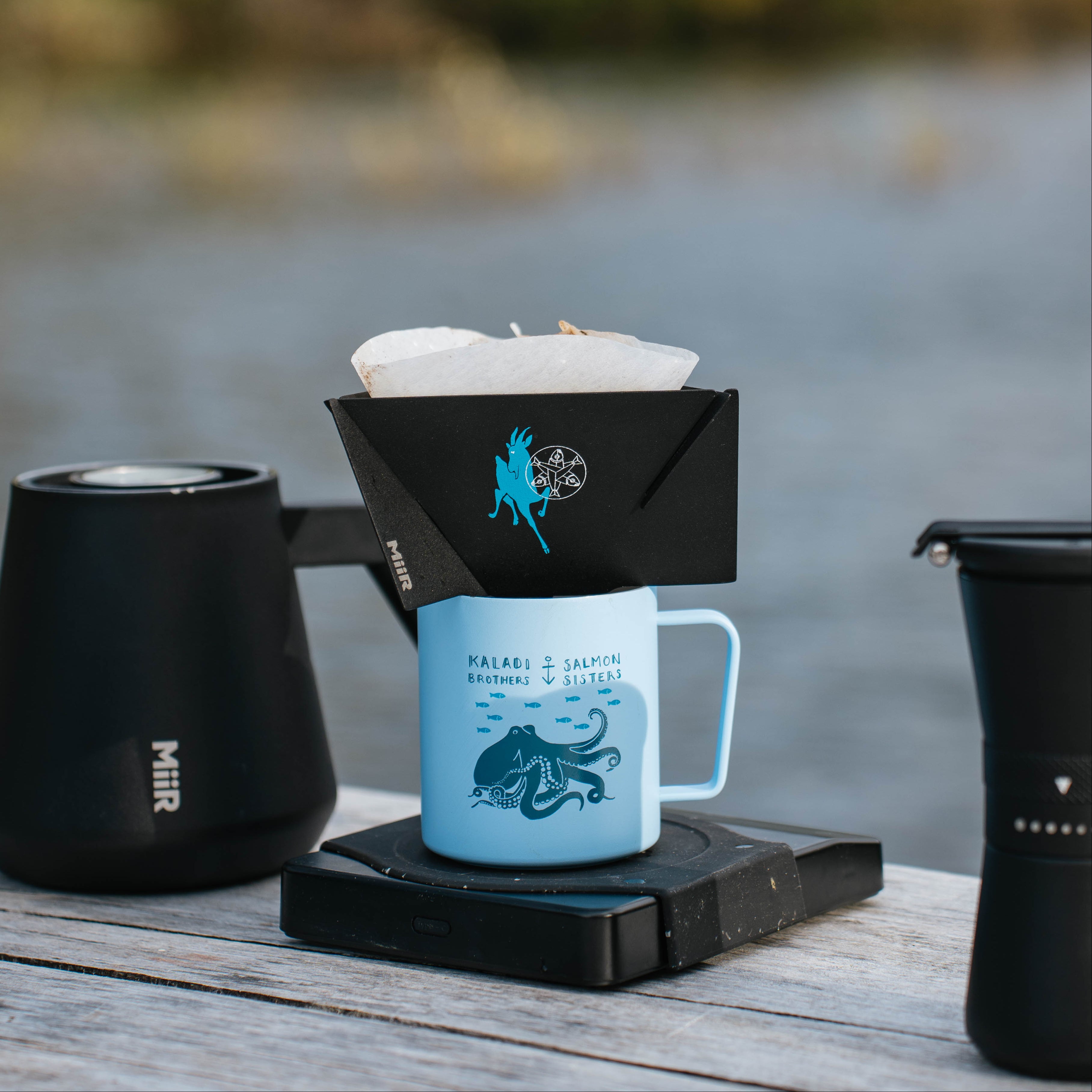 Coffee brewing setup with a blue mug, black filter holder, and black thermos on a wooden surface.