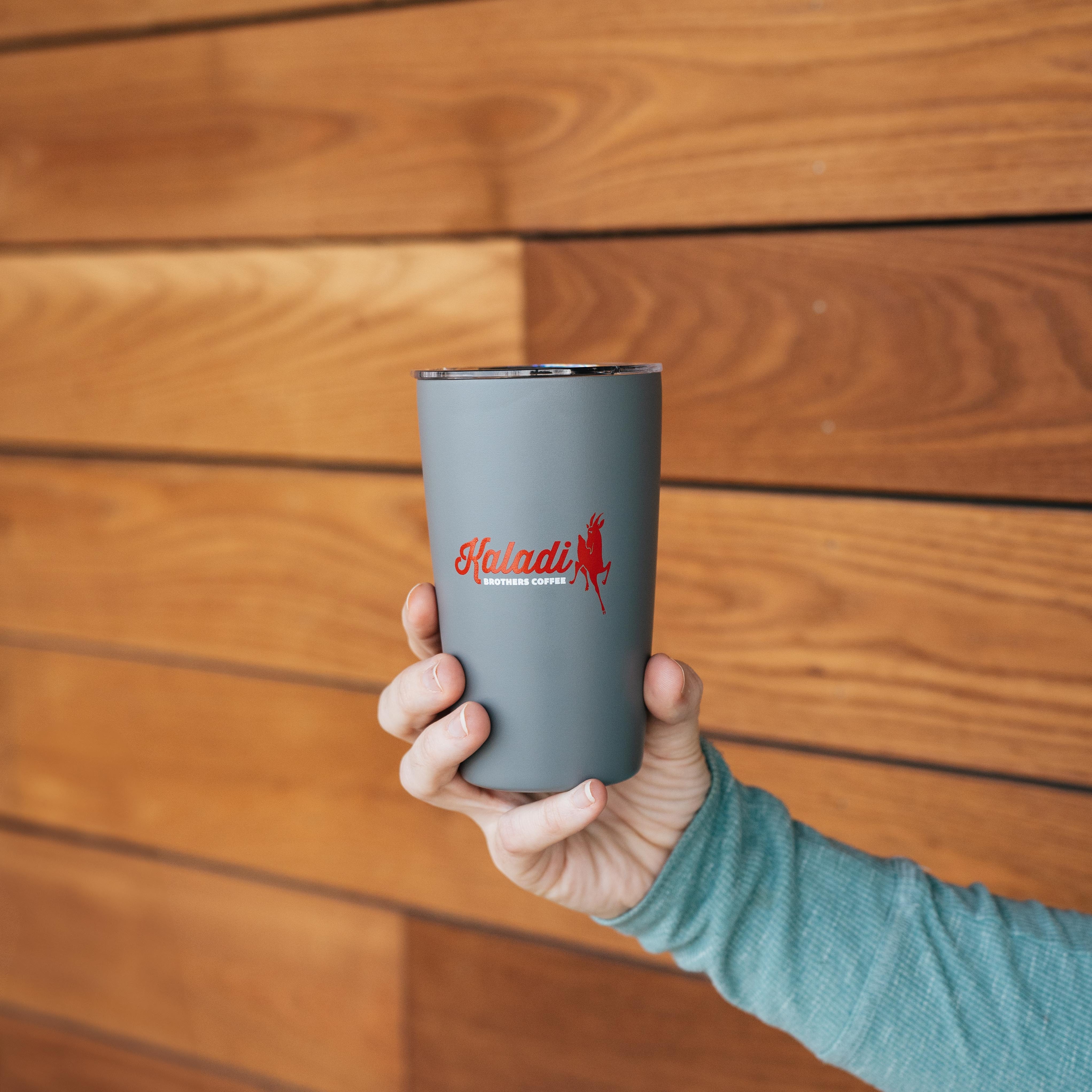 Hand holding the gray 12oz Kaladi tumbler against a wood-paneled wall