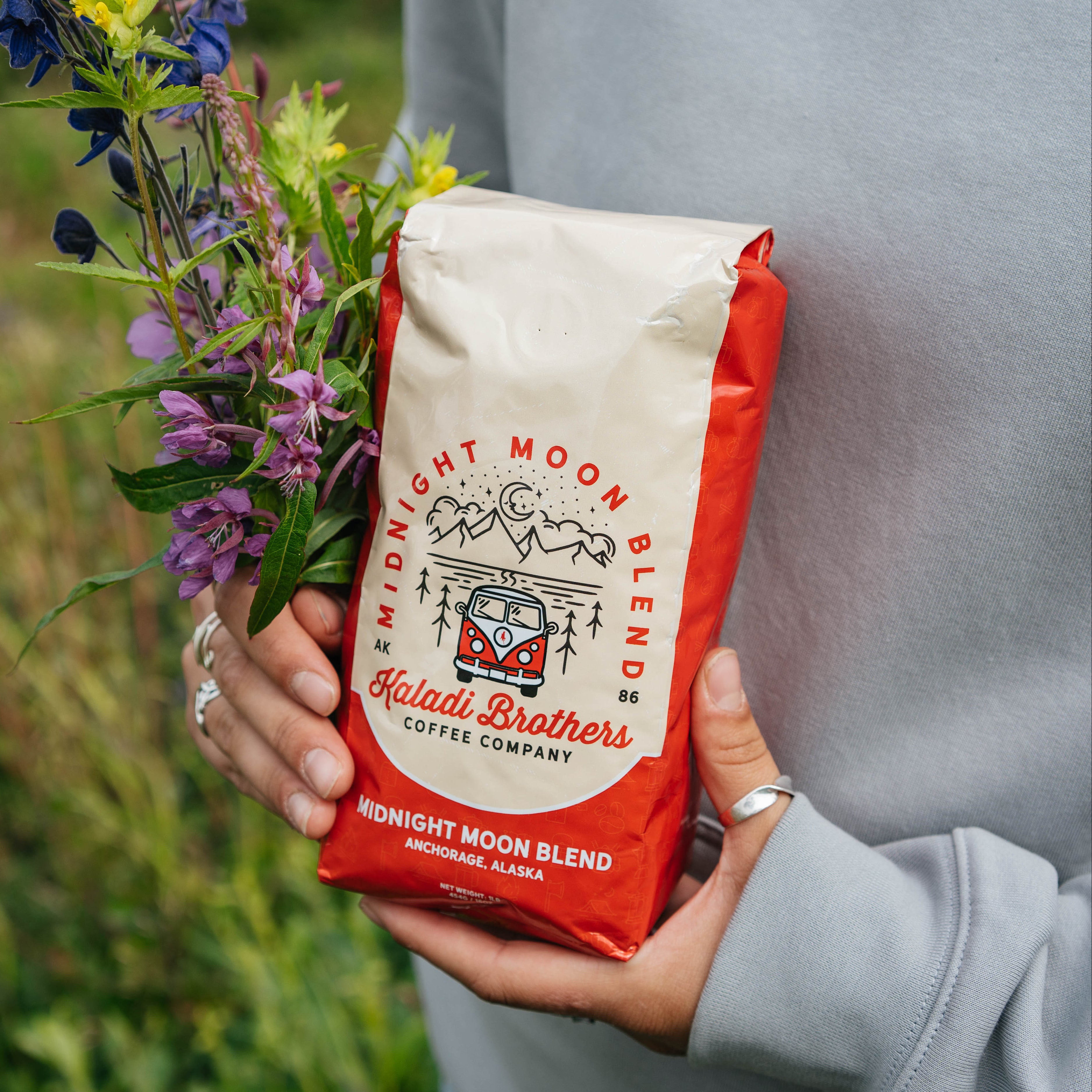Person holding a red Midnight Moon Blend coffee bag with a bouquet of wildflowers