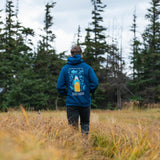 Person wearing a blue hoodie with a logo standing in a field with trees in the background