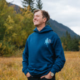 Man wearing the blue Mug Up Hoodie in an autumn field with mountains behind