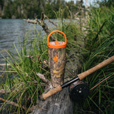A camouflage insulated tumbler with an orange lid and handle sits on a log beside a fishing rod at a lakeshore, with tall grass and water in the background.