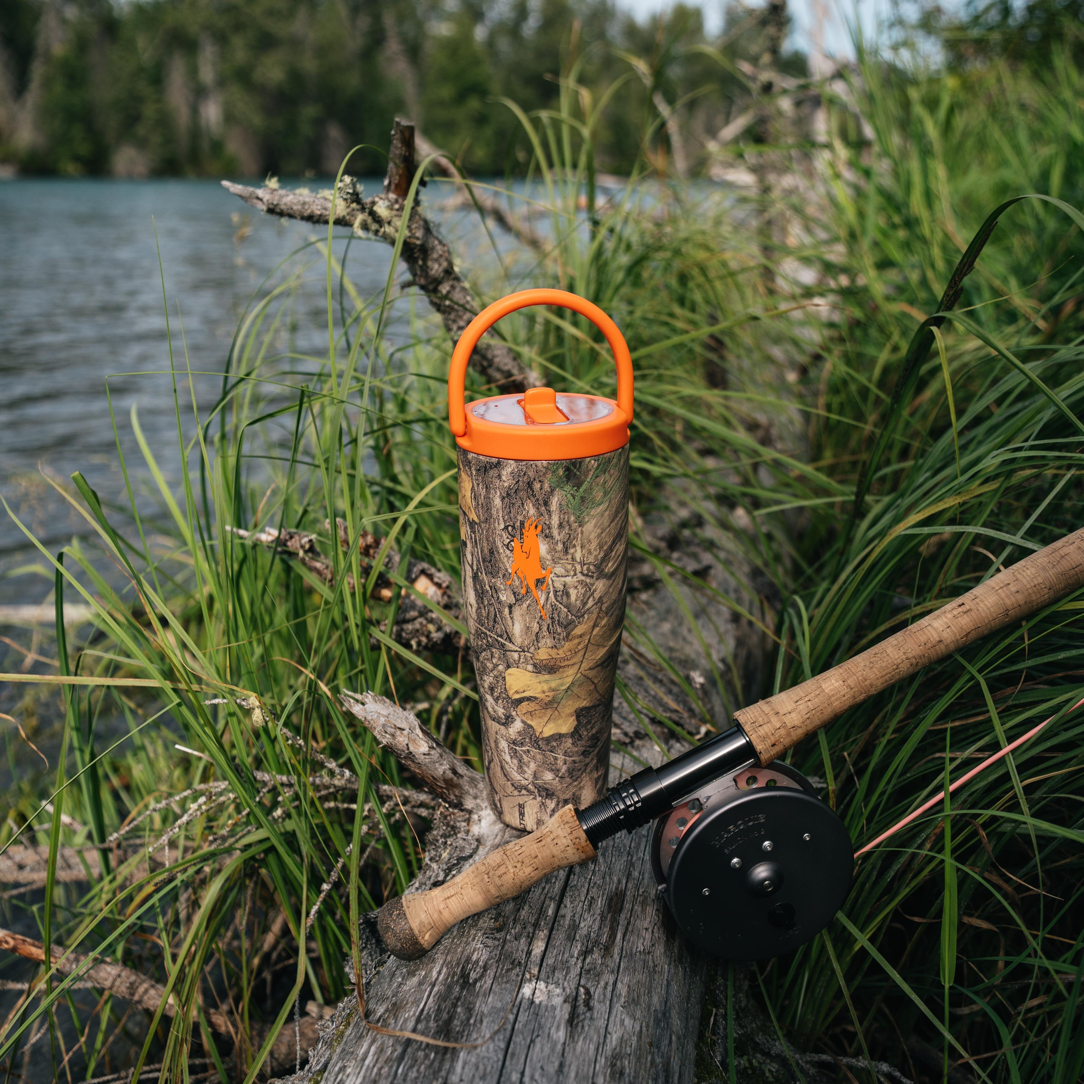 A camouflage insulated tumbler with an orange lid and handle sits on a log beside a fishing rod at a lakeshore, with tall grass and water in the background.