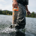A fly fisherman wading through shallow water while holding a camouflage insulated water bottle with a bright orange handle and lid.
