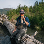 A man wearing fishing waders, a blue shirt, sunglasses, and a cap sits on a large fallen log in a river, holding an orange and white insulated bottle. The background features lush green trees and a distant mountain under a cloudy sky.