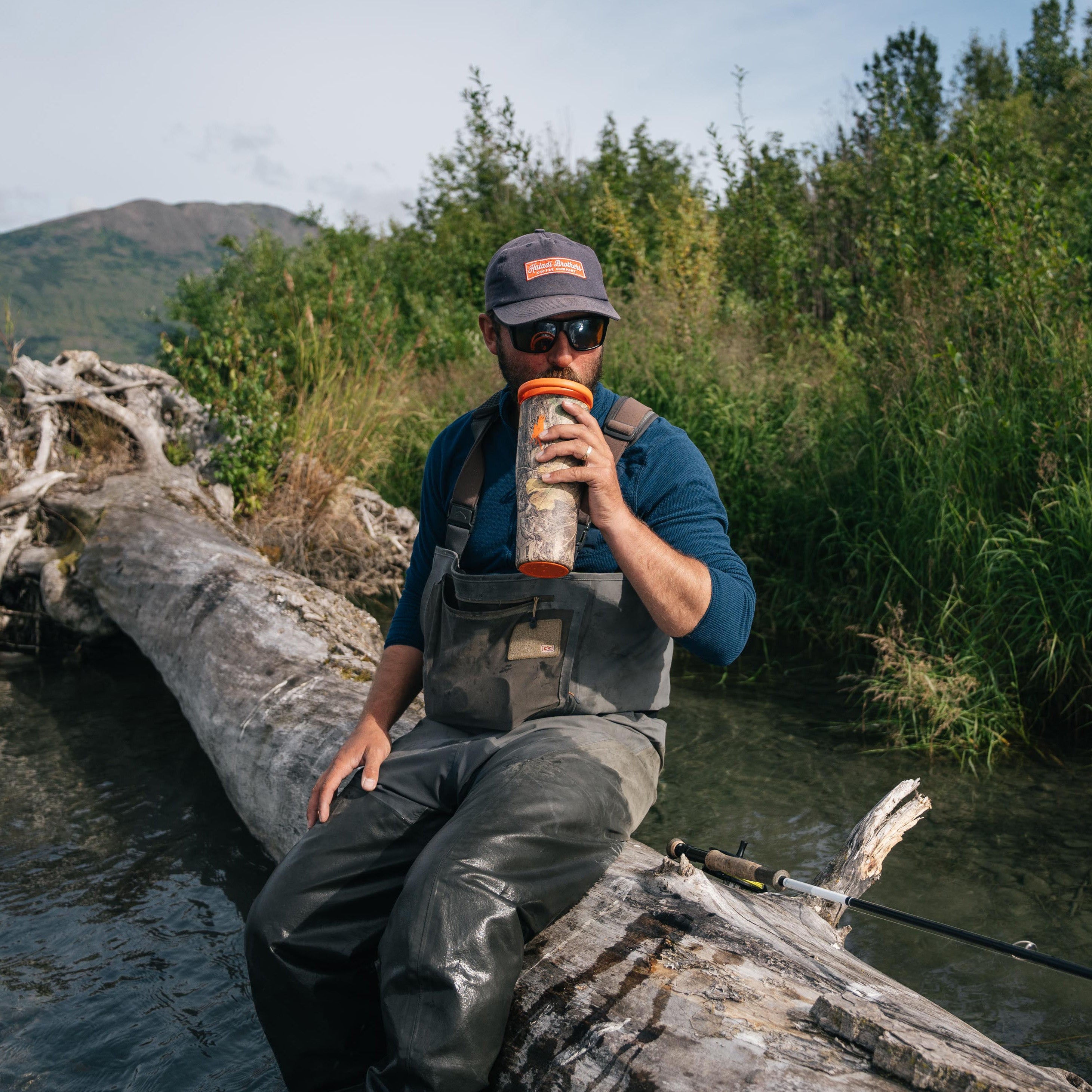 A man wearing fishing waders, a blue shirt, sunglasses, and a cap sits on a large fallen log in a river, holding an orange and white insulated bottle. The background features lush green trees and a distant mountain under a cloudy sky.